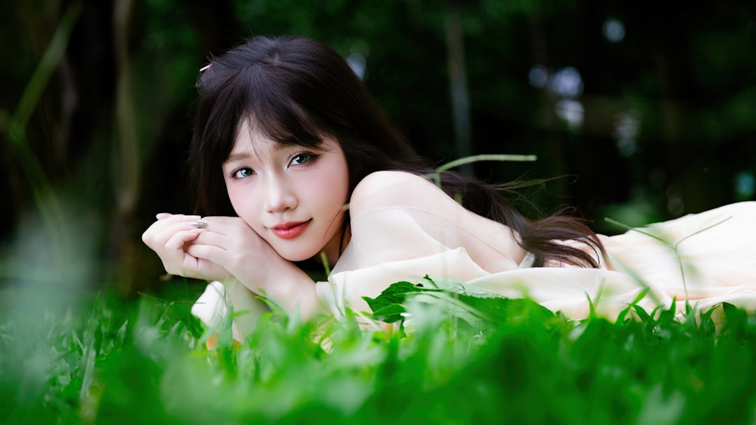 Smiling young woman lying on green grass with a bouquet of fresh hydrangea flowers, wearing a soft cream dress in natural light.