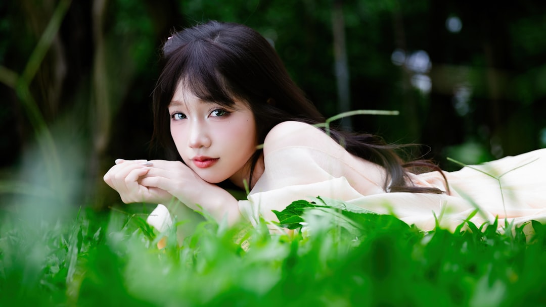 Smiling young woman lying on green grass with a bouquet of fresh hydrangea flowers, wearing a soft cream dress in natural light.